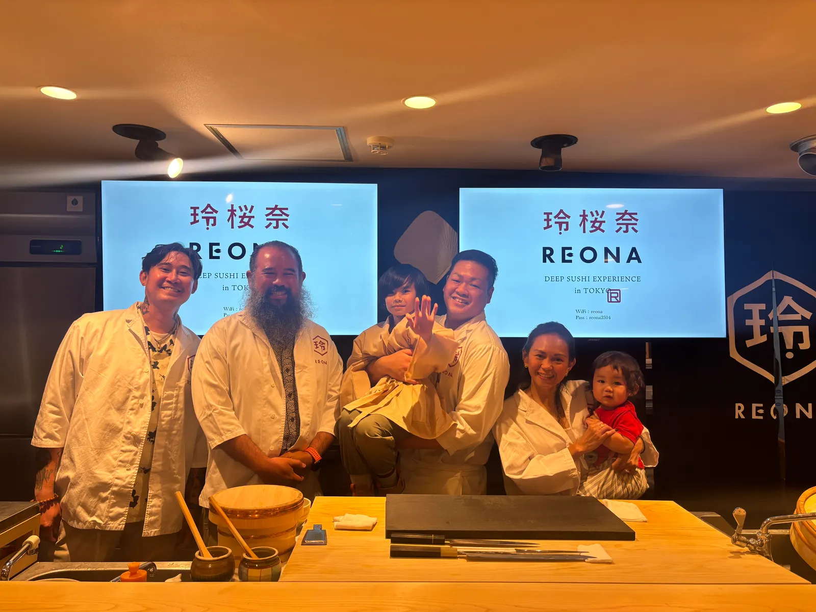 A family in REONA chef coats behind the sushi counter, with the chef holding a smiling child waving at the camera