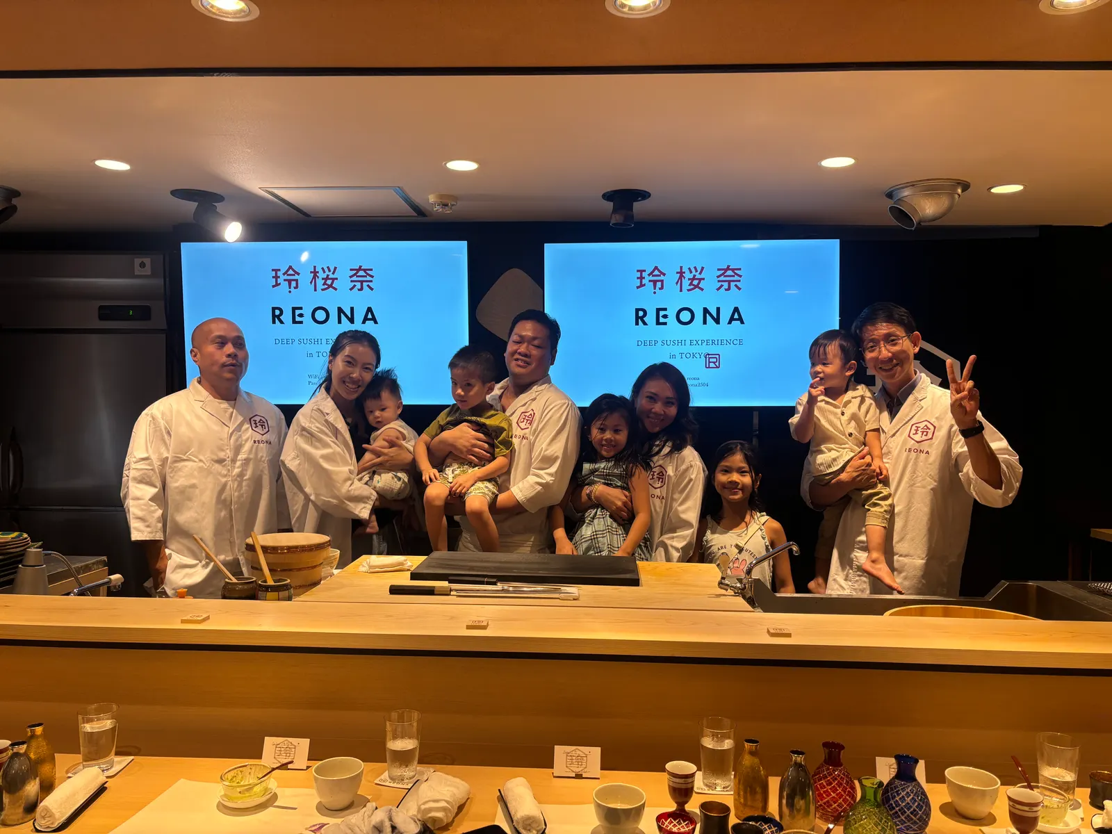 Two families with young children in REONA chef coats gathered behind the counter with the chef