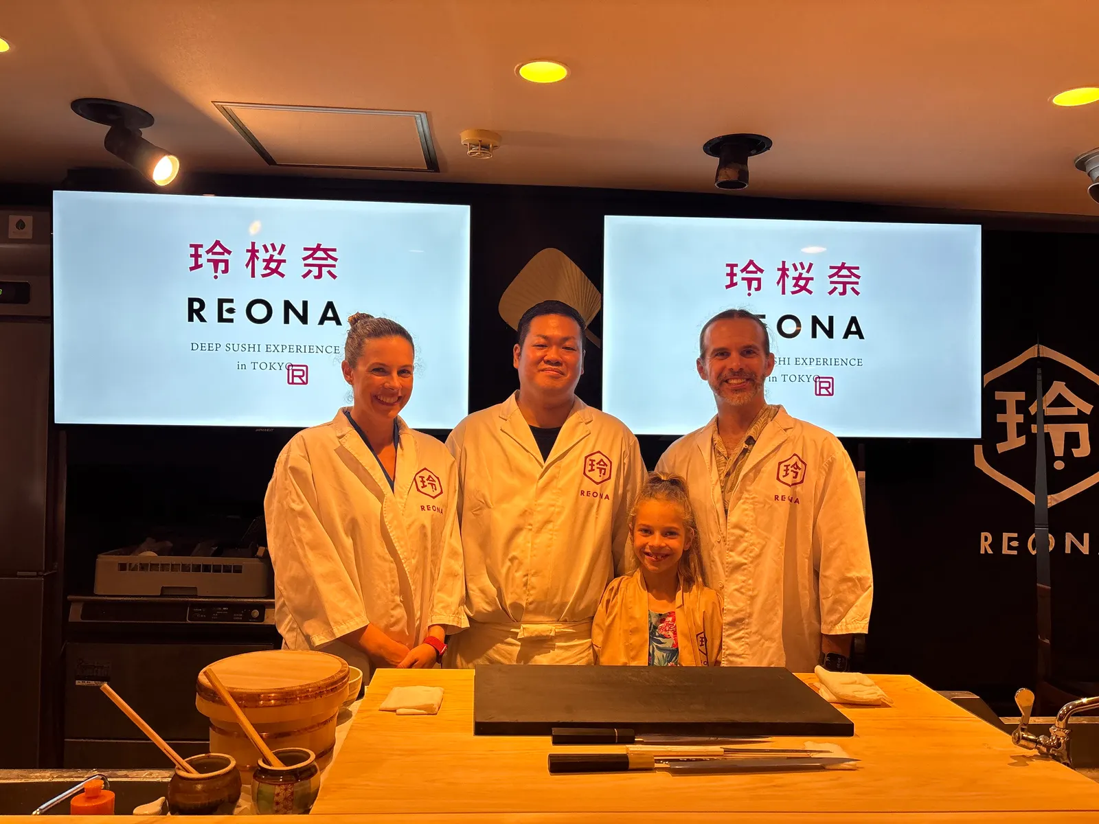 Parents and their young daughter in REONA chef coats posing with the chef behind the sushi counter