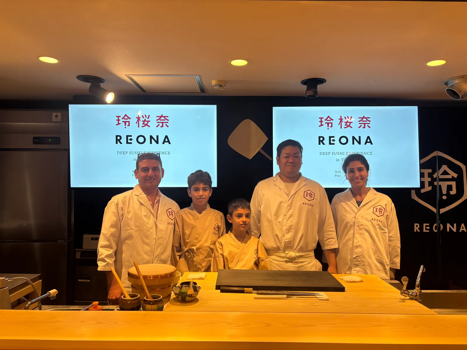 A family with two boys in REONA chef coats standing with the chef behind the counter
